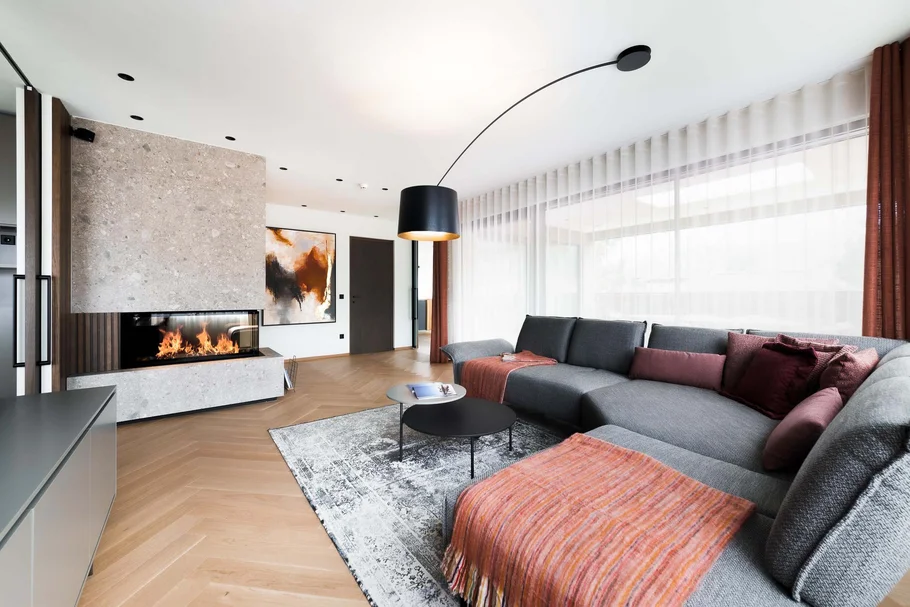 INTERIOS. Dein Interior-Design-Studio. Moderner Wohnzimmer mit grauem Sofa, Kamin und großen Fenstern mit weißen Vorhängen