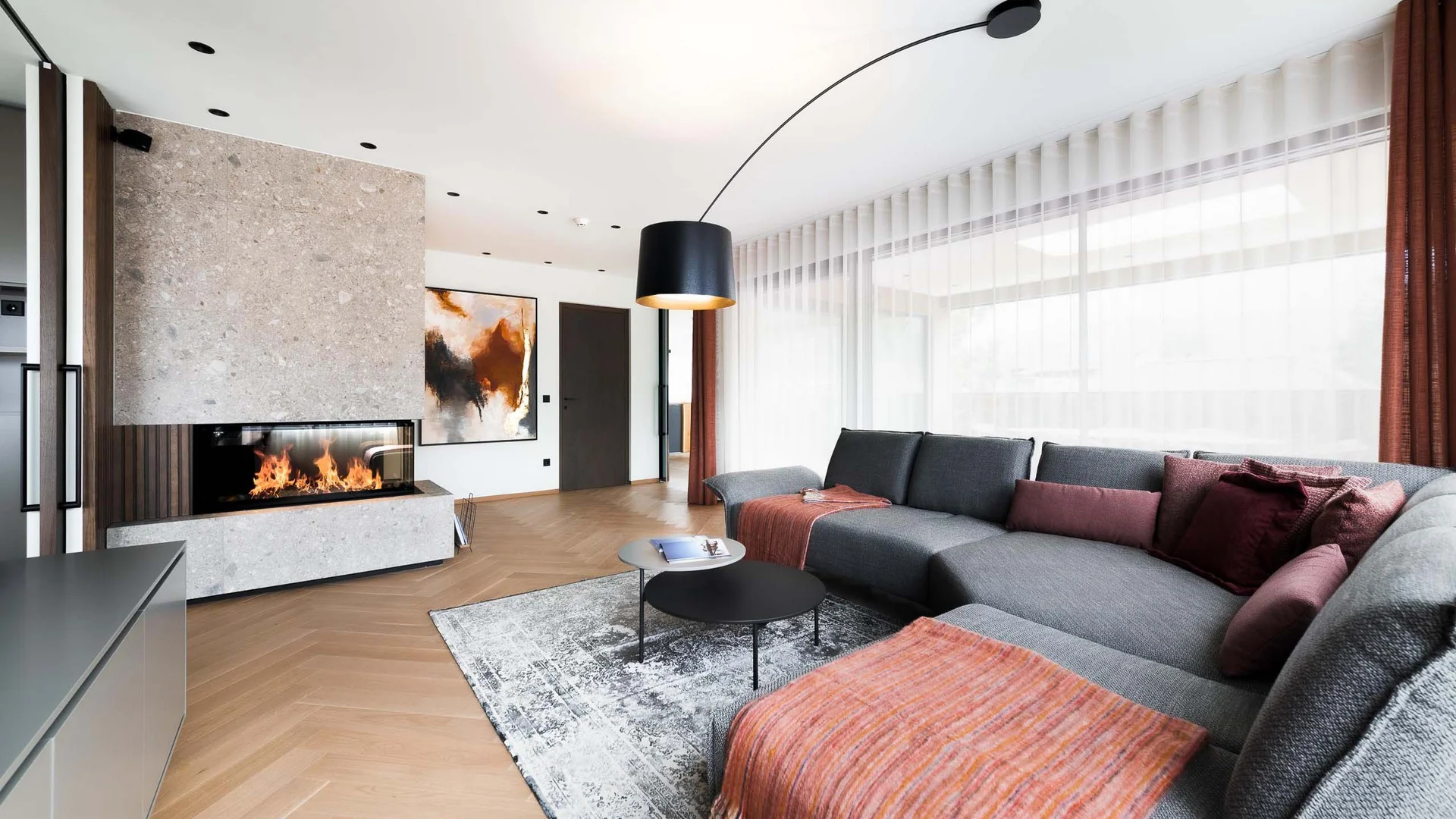 INTERIOS. Dein Interior-Design-Studio. Moderner Wohnzimmer mit grauem Sofa, Kamin und großen Fenstern mit weißen Vorhängen