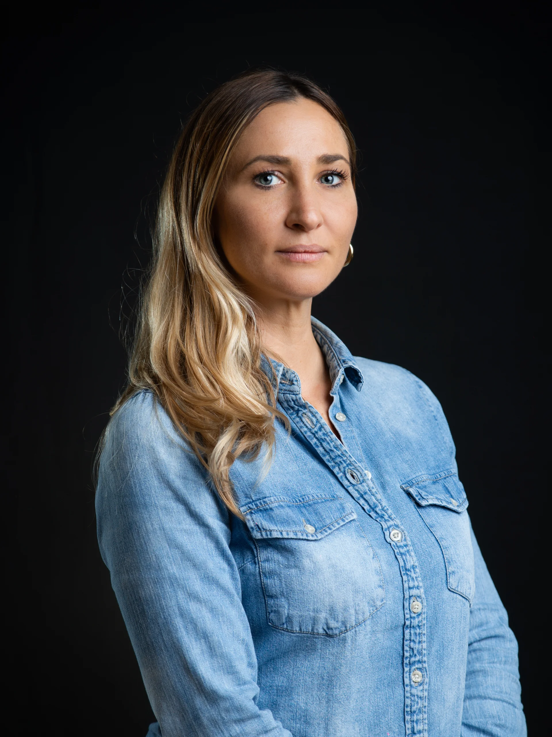 INTERIOS. Dein Interior-Design-Studio. Frau mit langem blonden Haar und Jeanshemd vor schwarzem Hintergrund