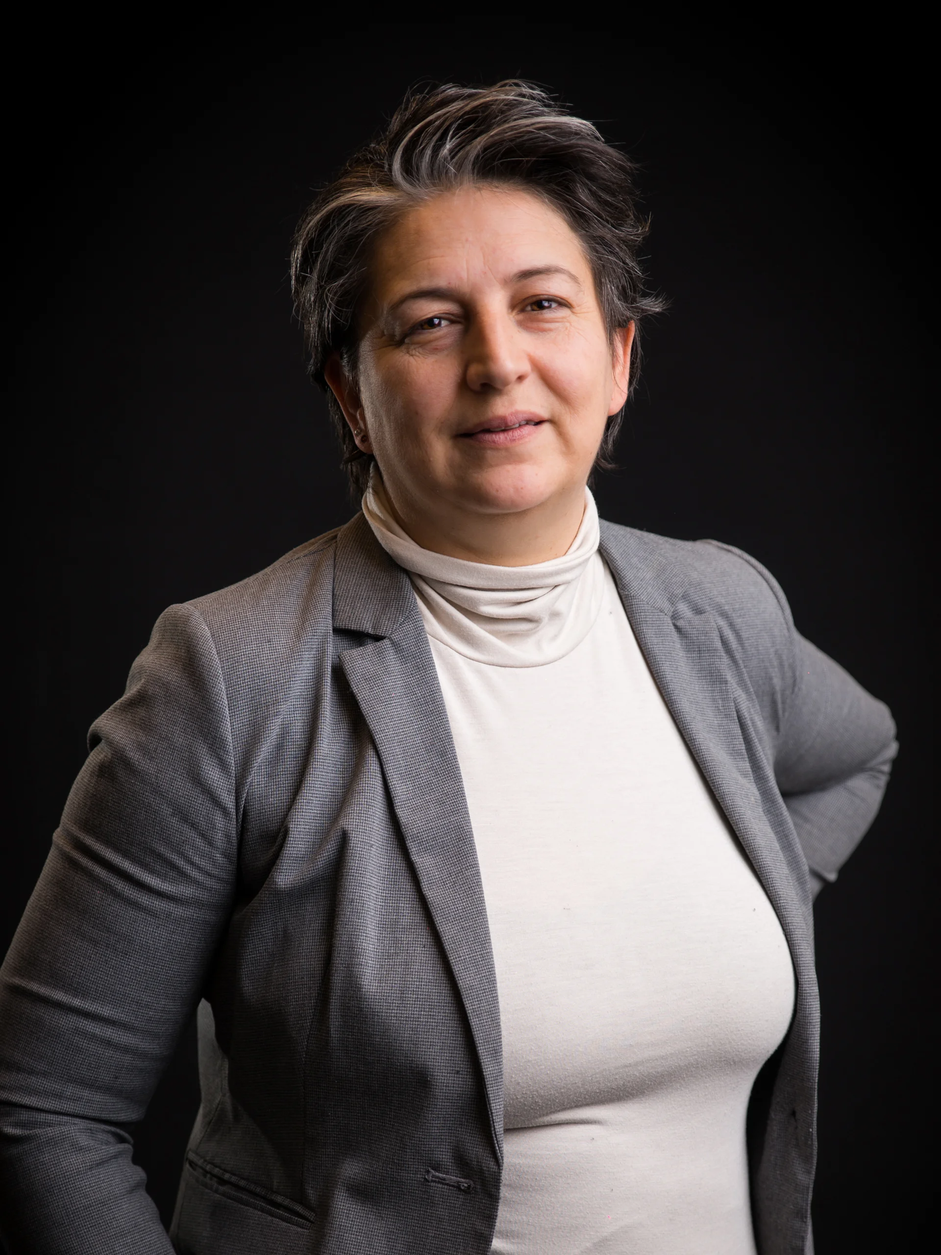 INTERIOS. Dein Interior-Design-Studio. Porträt einer Frau mit kurzem dunklem Haar, grauem Blazer und hellem Oberteil vor schwarzem Hintergrund