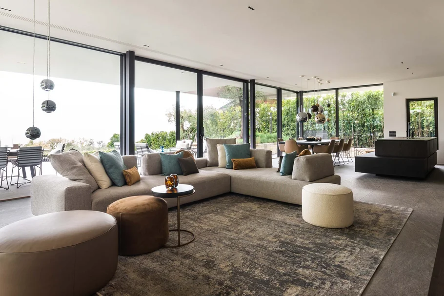 INTERIOS. Dein Interior-Design-Studio. Moderner Wohnbereich mit Ecksofa, Sitzhockern und großen Glasfenstern