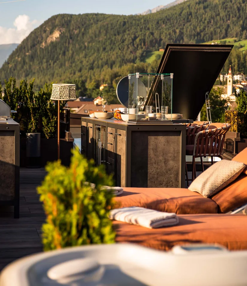 INTERIOS. Dein Interior-Design-Studio. Sonnige Dachterrasse mit Liegestühlen, Außengrill und Bergblick