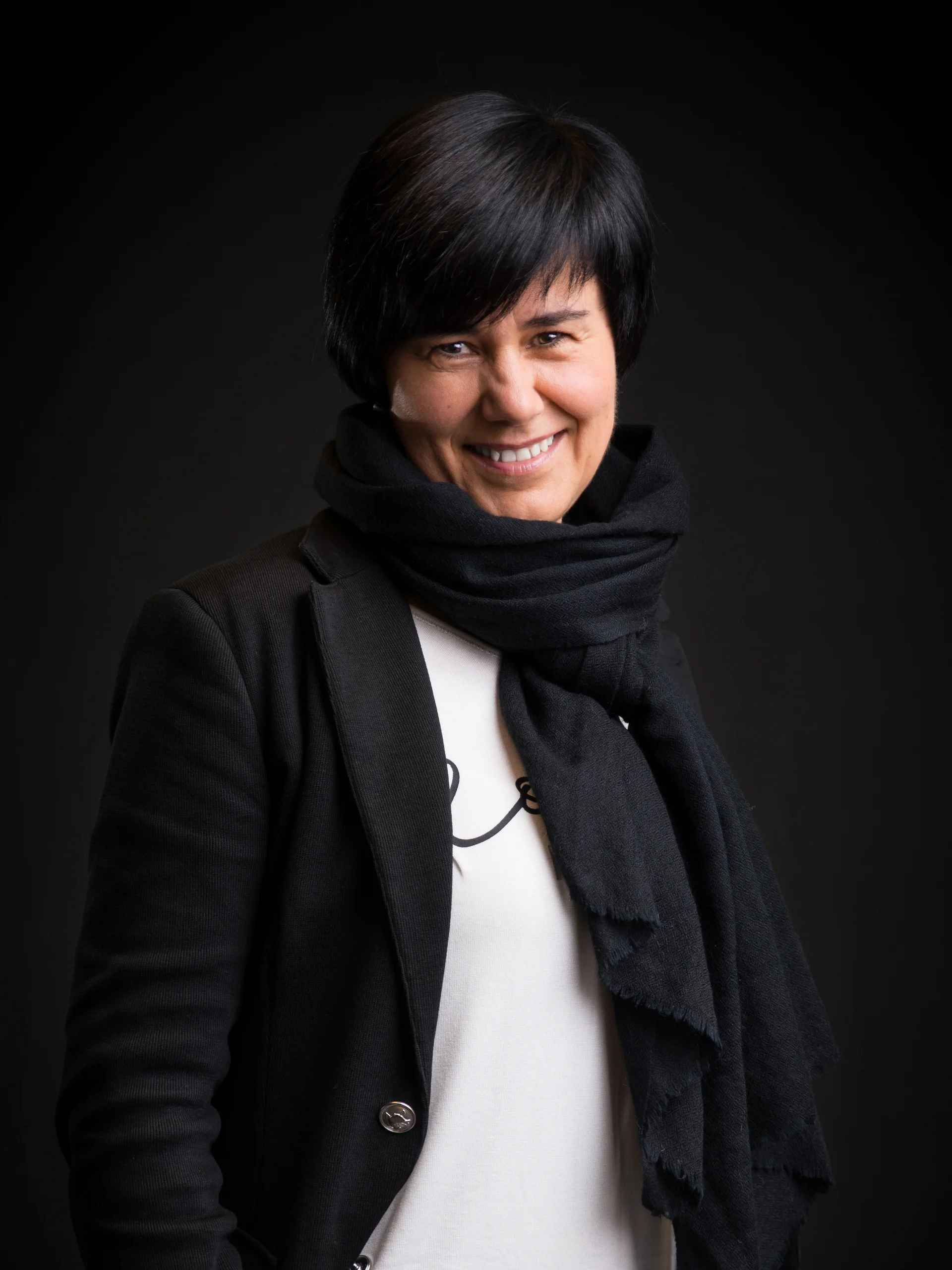 INTERIOS. Dein Interior-Design-Studio. Lächelnde Person mit schwarzem Mantel und schwarzem Schal vor dunklem Hintergrund