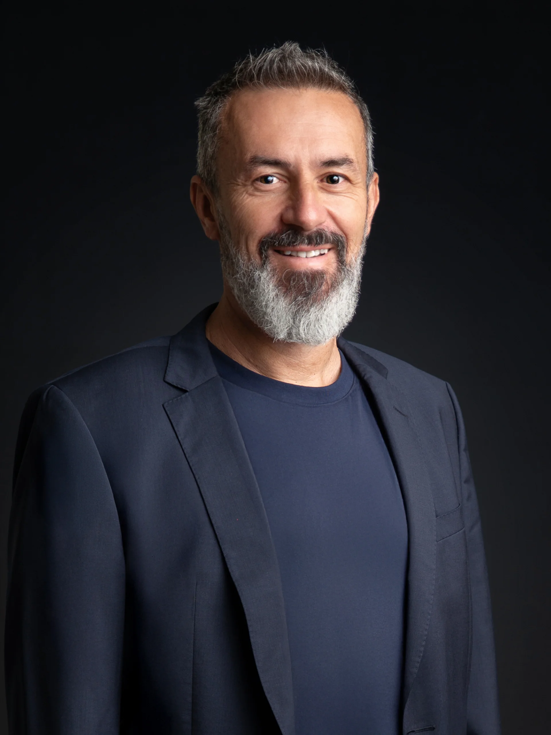 INTERIOS. Dein Interior-Design-Studio. Lächelnder Mann mit grauem Bart in dunklem Anzug vor schwarzem Hintergrund