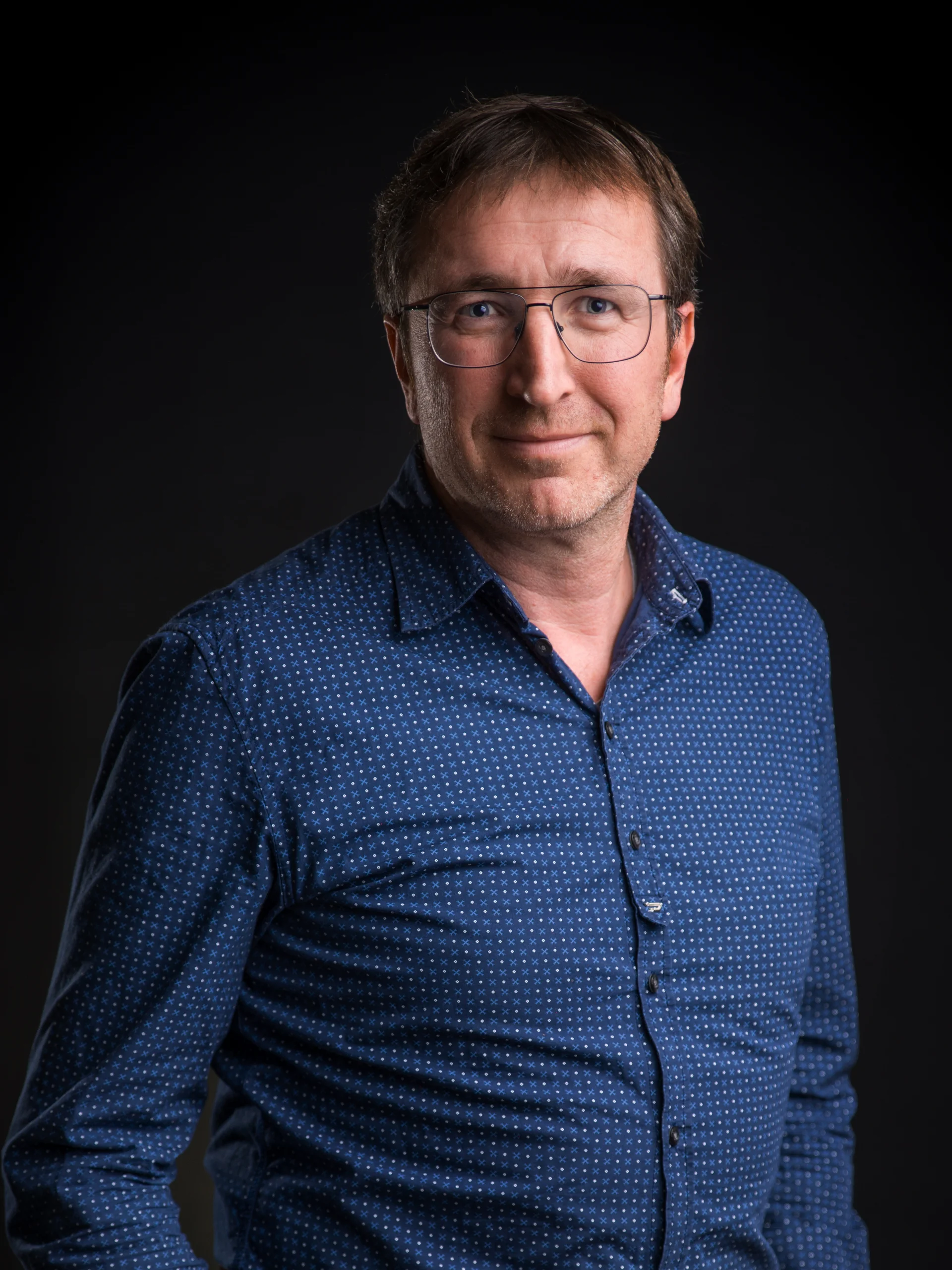 INTERIOS. Dein Interior-Design-Studio. Mann mit Brille und blauem Hemd vor schwarzem Hintergrund