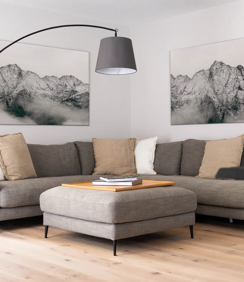 INTERIOS. Dein Interior-Design-Studio. Gemütliche Sitzecke mit grauem Ecksofa, Hocker, Stehlampe und Bergbildern
