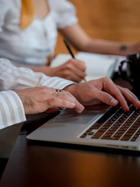 Marketing- und Verkaufsförderung Person tippt auf Laptop, andere schreibt im Hintergrund in Notizbuch
