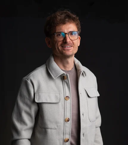 INTERIOS. Dein Interior-Design-Studio. Mann mit Brille trägt hellgraue Jacke vor schwarzem Hintergrund