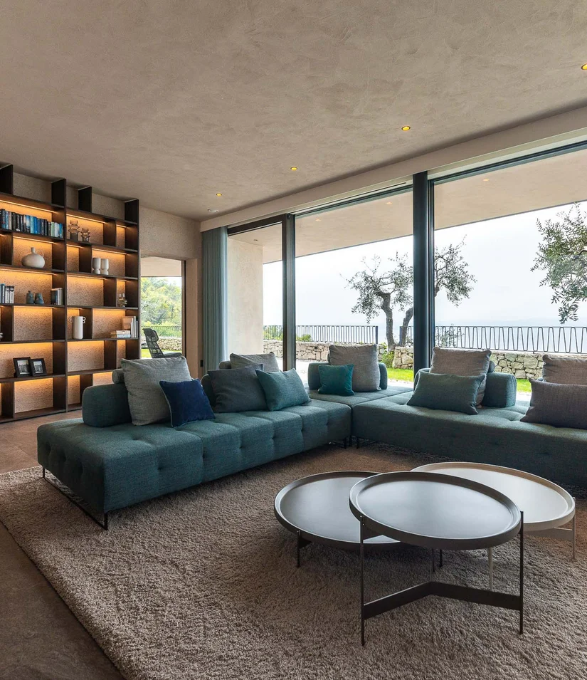 INTERIOS. Dein Interior-Design-Studio. Moderner Wohnbereich mit großem Sofa und Blick auf Terrasse mit Bäumen