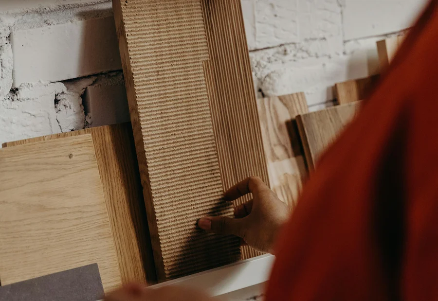 INTERIOS. Dein Interior-Design-Studio. Person wählt Holzplattenmuster von einem Regal an weißer Backsteinwand