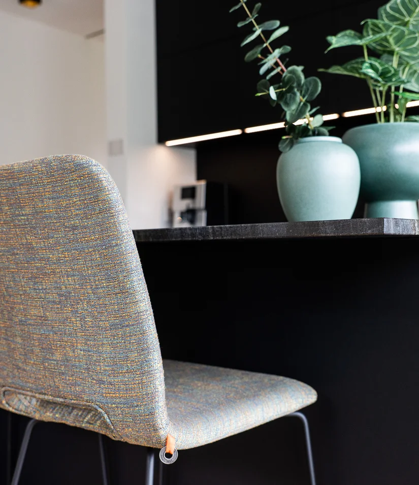 INTERIOS. Dein Interior-Design-Studio. Zwei gepolsterte Stühle an einer schwarzen Kücheninsel mit Dekopflanzen