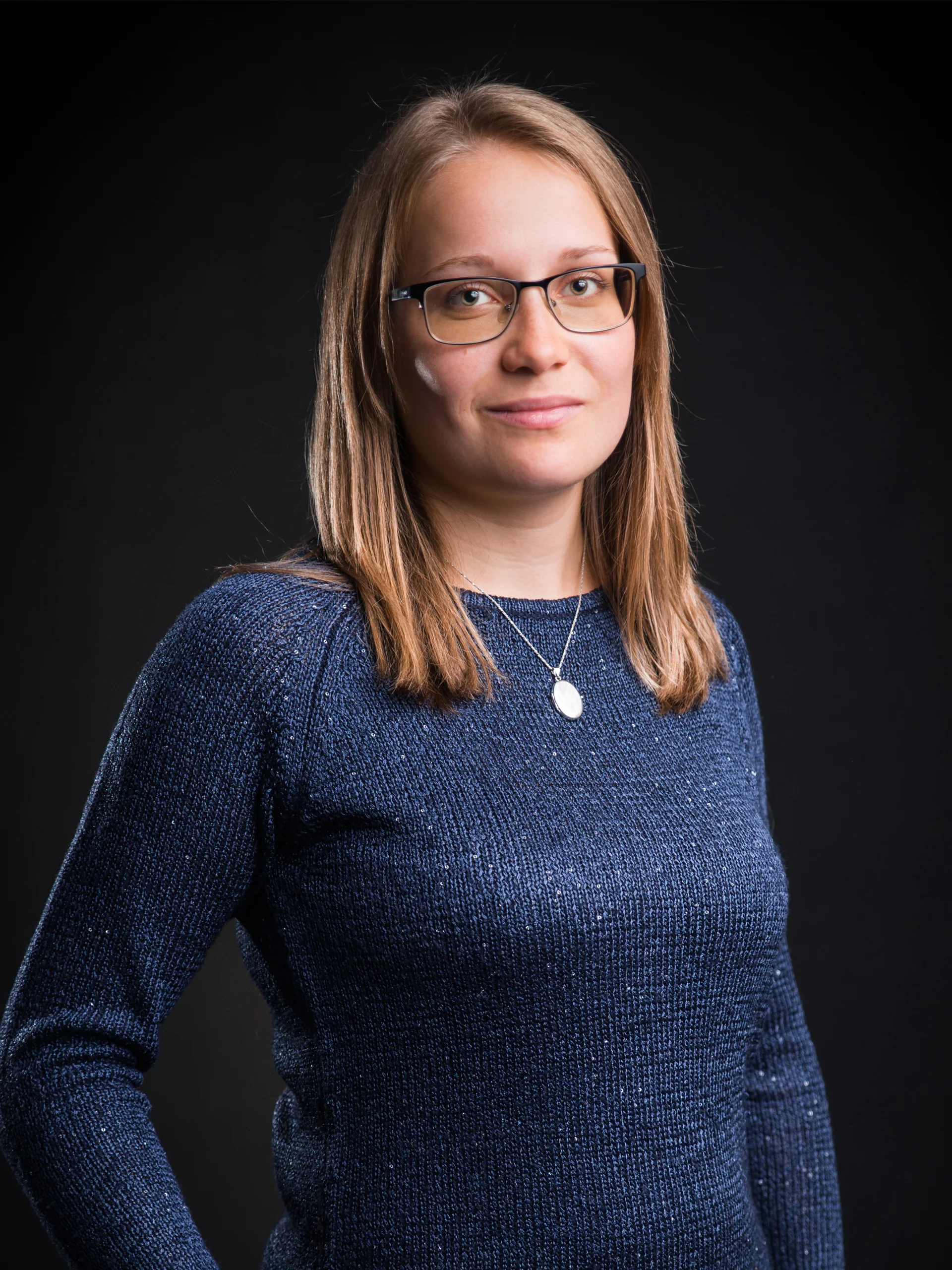 INTERIOS. Dein Interior-Design-Studio. Frau mit Brille und blauem Pullover vor schwarzem Hintergrund